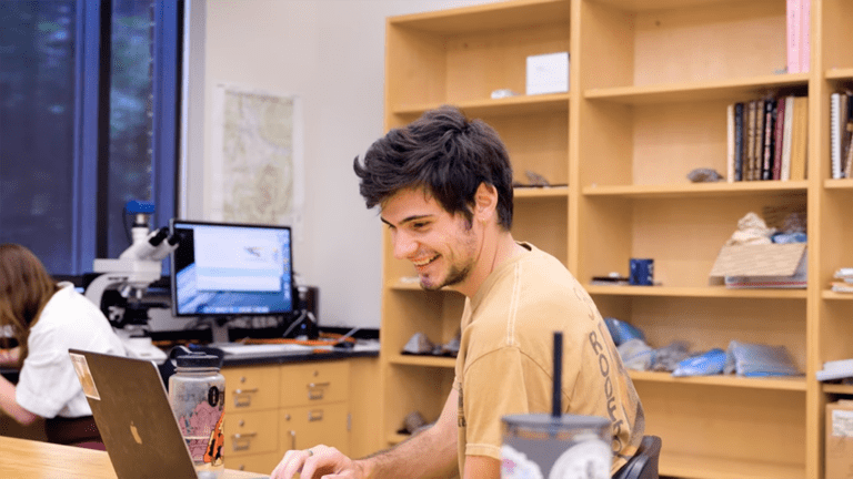 A young man with dark hair sits at a lab bench with a microscope in front of him. He's looking to the side and laughing. 